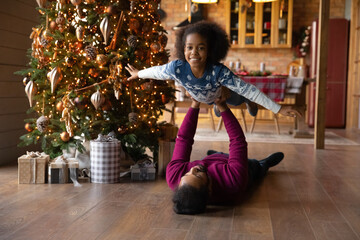 Wall Mural - Portrait African American father lying on warm floor, carrying smiling adorable daughter, celebrating Christmas at home, loving dad and little girl playing active game, having fun on winter holiday