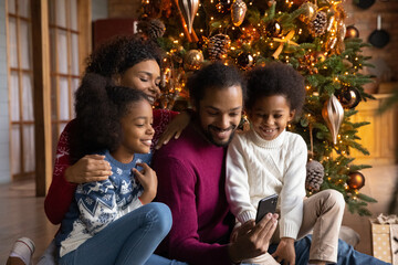 Wall Mural - Happy African American family with two kids using smartphone, sitting near festive tree at home, smiling mother and father with son and daughter chatting or shopping online on Christmas holiday