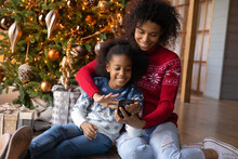 Happy African American Woman With Adorable Little Daughter Using Phone, Looking On Smartphone Screen, Shopping Online, Choosing Gifts, Sitting On Warm Floor Near Christmas Tree At Home