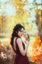 Bright Autumn Fairy Tale. Fantasy Woman Hugs A Barn Owl. Forest Nymph Girl Holds A White Bird In Her Hands. Portrait Of Romantic Lady In A Creative Dress. Background Of Nature, Orange Yellow Trees