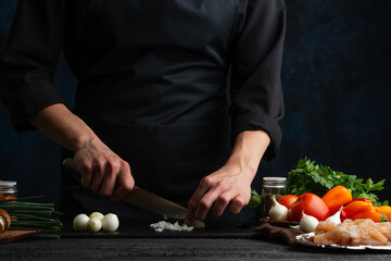 Wall Mural - The chef in black uniform cuts with knife garlic n black chopped board at the professional kitchen with ingredients on dark background.