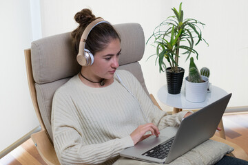 Wall Mural - High angle shot of a young European woman using a laptop with headset sitting on the chair at home