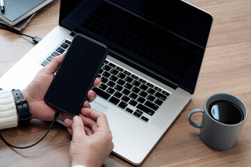 Wall Mural - Cropped shot of man hand holding black screen smartphone while sitting at the table with laptop and cup of coffee.