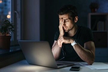 Young businessman has headache while working overtime with laptop at home office late night. Stressed depressed freelancer touching his head, feeling pain in eyes. Insomnia, internet addiction