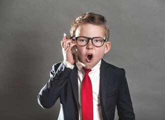 Angry child boss screaming on the mobile phone. School boy in suit and tie and glasses as a businessman in the office