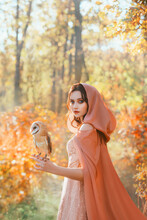 Portrait Of Fantasy Girl Princess Walks With White Bird Barn Owl On Her Hand. Beautiful Mystical Woman In Medieval Cape Cloak With Hood Looks Into Camera. Background Bright Autumn Nature, Forest Trees