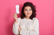 Good looking feminine girl holds clean sanitary napkin, has menstrual cycle, happy to have good women health, female with dark wavy hair wearing white warm sweater isolated over pink background.