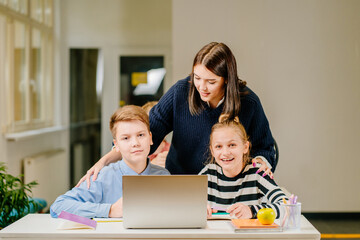 Canvas Print - Two happy schoolchild pupils boy and girl woth their tutor working together at lesson in classroom. Female teacher helping explain the task for her two students