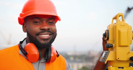 Wall Mural - Portrait of cheerful handsome young African American man in casque turning to camera at construction site and smiling at total station. Good looking male builder at building outdoor. Construct concept