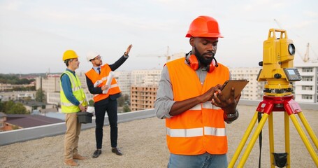 Wall Mural - African American man topographer in casque measuring angle with total station on roof of building. Male builder constructor doing topographic measures and using tablet device. Geodesy concept Geodesic