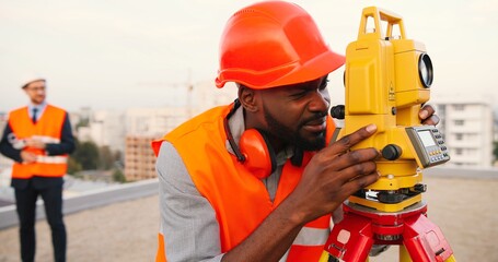 Wall Mural - African American young man topographer in casque measuring angle with total station on roof of building. Male builder. Constructor doing topographic measures. Geodesy concept. Geodesy. Constructing.