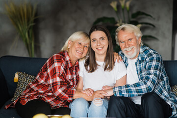 Family portrait of a Caucasian family. Pretty daughter is sitting on the couch between her elderly beloved parents, they look at the camera and smiling