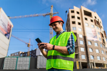 Portrait of young engineer man use phone while standing at construction area. Builder in helmet control according to plan by mobile phone.