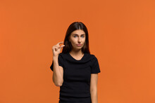 A Little Bit More. Portrait Of Beautiful Caucasian Woman In Black T-shirt Forms Something Tiny, Makes Hand Gesture, Has Beg Facial Expression. Isolated Over An Orange Background