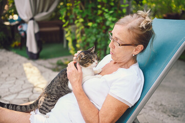 Wall Mural - Happy smiling senior woman in glasses relaxing in summer garden outdoors hugging domestic tabby cat