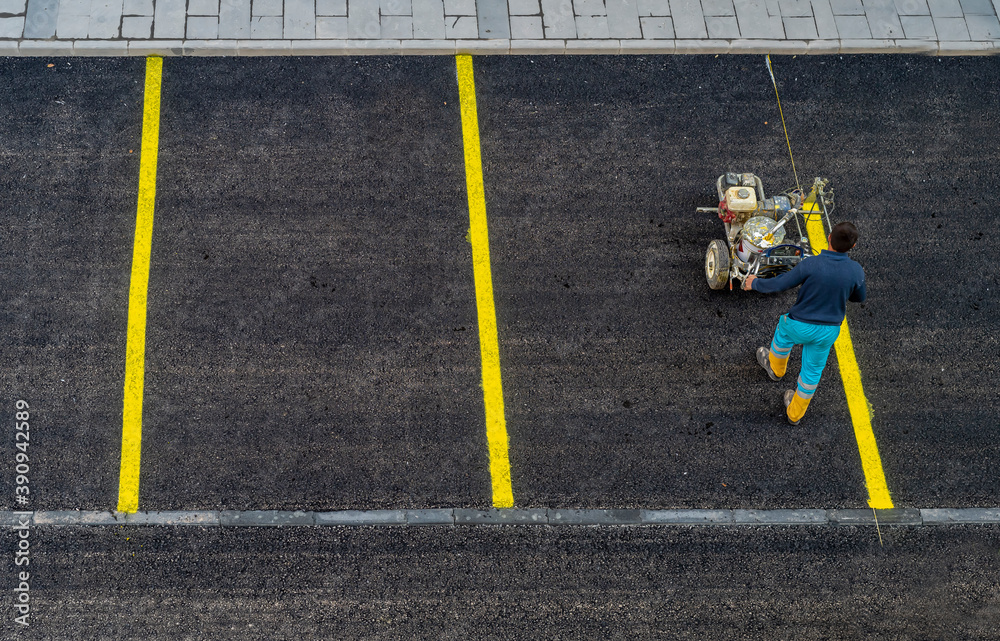 A worker is drawing outdoor concrete car parking lot is divided for making a slot for the park each car by a yellow line, with cement wheel coverings. in urban city. - obrazy, fototapety, plakaty 