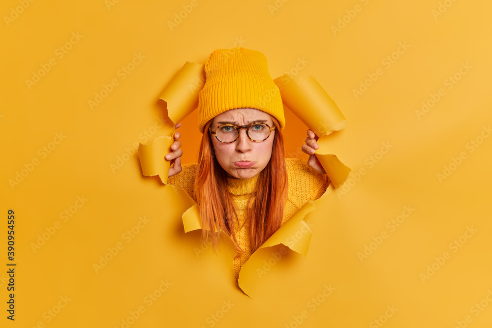 Unhappy disappointed young woman purses lips and looks sadly at camera stands offended breaks through paper background against yellow background. Upset gloomy redhead teenage girl in spectacles - obrazy, fototapety, plakaty 