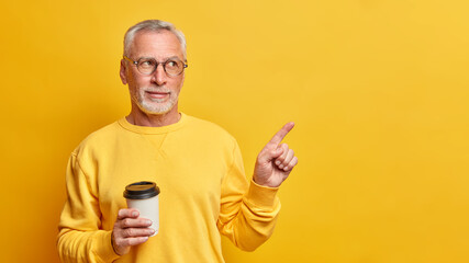 Wall Mural - Serious wrinkled grey haired man drinks aromatic takeaway coffee indicates at empty space and shows place for your advertising content dressed in casual jumper isolated over yellow background