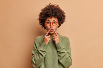 Lovely dark skinned young woman keeps index fingers near folded lips makes face at camera wants to kiss someone wears glasses and sweater isolated over brown background. Face expressions concept