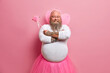 Cheerful confident bearded bald man fairy going to make your wish come true holds magic wand wears wings and crown has big abdomen isolated over pink studio background. Holiday performer indoor