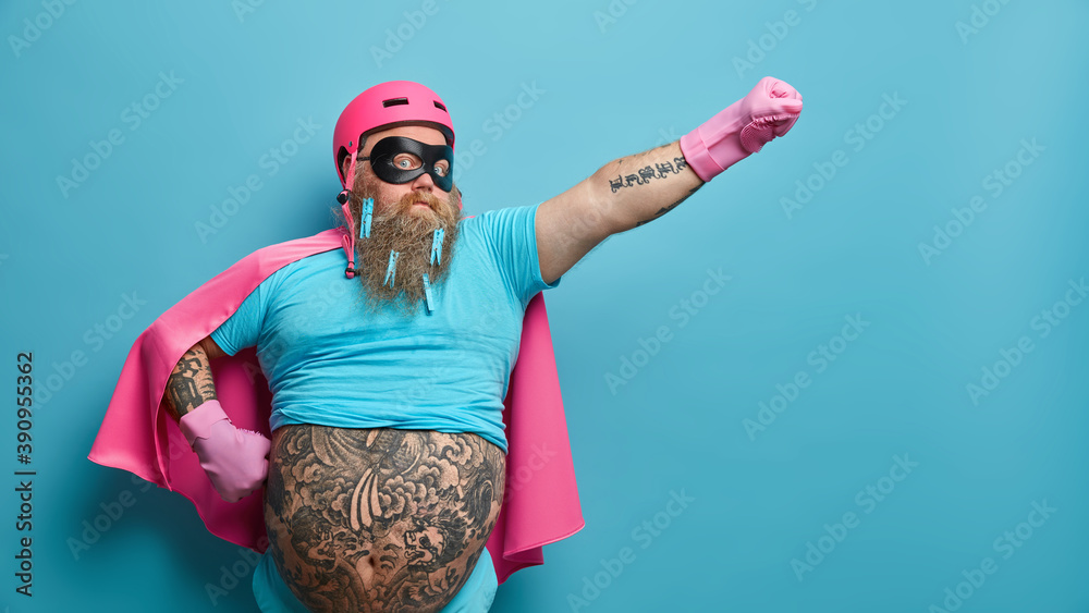 Self confident serious bearded man ready to help people wears superhero costume pretends being heroic character has supernatural power stretches arm over blue background with empty blank space - obrazy, fototapety, plakaty 