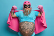 © wayhome.studio  - Serious bearded brave super natural man raises arms and shows muscles demonstrates his power has big tattooed stomach wears pink helmet cape rubber gloves and mask poses over blue studio wall
