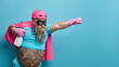 Horizontal shot of self confident male hero wears pink helmet cloak and rubber gloves holds bottle of detergent ready to help you with cleaning stretches arm as going to fly isolated over blue wall