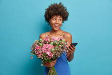 Positive Dark Skinned Woman With Curly Hair Holds Nice Bouquet Of Flowers Gets Order Online Uses Modern Smartphone Enjoys Present From Boyfriend Looks Aside With Smile Isolated On Blue Wall.