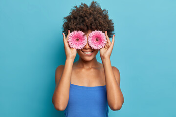 Curly haired woman holds flowers over eyes bites lips and hides face has summer mood dressed in blue clothing in one tone with background. Florist going to make bouquet from gerbera daisies.