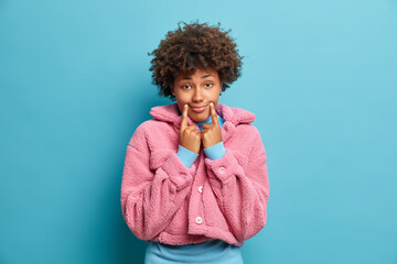 Wall Mural - Sad dark skinned woman tries to be happy keeps finger near corners of lips forces smile looks with gloomy expression at camera wears pink fur coat isolated over blue background. Negative emotions