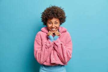 Wall Mural - Tender pleased dark skinned young woman smiles lovely keeps hands under chin dressed in pink winter coat has curly hair poses against blue background. Positive face expressions fashion concept