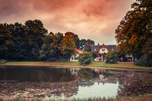 Rural Buildings By A Reflective Pond With Fallen Autumn Foliage On The Water Surface In Burgerpark