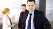 © cameravit - Portrait of a self-confident middle aged businessman in a blue suit, standing in a sunny modern office with his colleagues at the background. Concept of business success