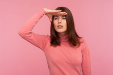 Curious Brunette Woman In Pink Sweater Looking Far Away With Hand Over Head, Trying To See Something, Bad Vision. Indoor Studio Shot Isolated On Pink Background