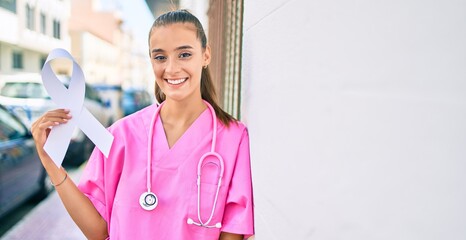 Wall Mural - Young doctor woman holding white cancer ribbon leaning on the wall at street of city.