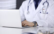 © rogerphoto - Unknown male doctor sitting and working with laptop in clinic at his working place, close-up. Young physician at work. Perfect medical service, medicine concept