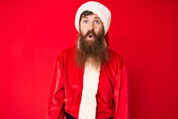 Wall Mural - Handsome young red head man with long beard wearing santa claus costume scared and amazed with open mouth for surprise, disbelief face