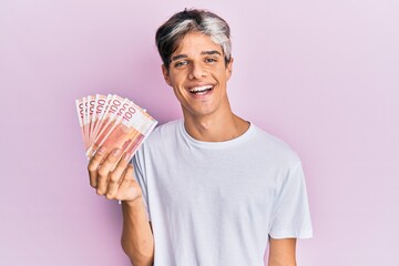 Sticker - Young hispanic man holding 100 norwegian krone banknotes looking positive and happy standing and smiling with a confident smile showing teeth