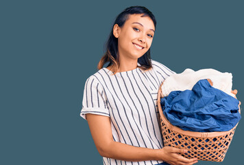 Sticker - Young woman holding laundry basket looking positive and happy standing and smiling with a confident smile showing teeth