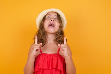 Wall Mural - young Caucasian girl standing against yellow background being amazed and surprised looking and pointing up with fingers showing something strange.