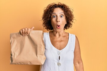 Sticker - Beautiful middle age mature woman holding take away paper bag scared and amazed with open mouth for surprise, disbelief face