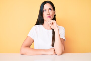 Sticker - Young caucasian woman sitting at the desk wearing casual white tshirt thinking concentrated about doubt with finger on chin and looking up wondering