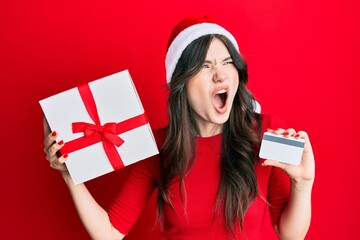 Sticker - Young beautiful caucasian girl wearing christmas hat, holding a gift and credit card angry and mad screaming frustrated and furious, shouting with anger. rage and aggressive concept.