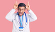 Young hispanic man wearing doctor uniform and stethoscope posing funny and crazy with fingers on head as bunny ears, smiling cheerful