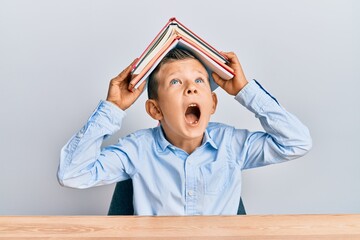 Wall Mural - Adorable caucasian kid holding book on head angry and mad screaming frustrated and furious, shouting with anger looking up.