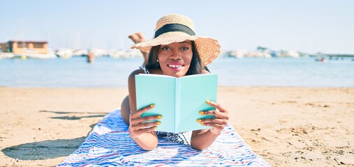 Young african american woman on vacation laying on the towel reading book at the beach