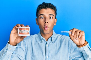 Sticker - Young handsome african american man holding invisible aligner orthodontic and braces making fish face with mouth and squinting eyes, crazy and comical.