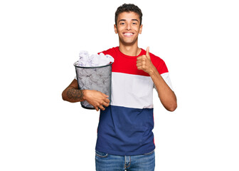 Wall Mural - Young handsome african american man holding paper bin full of crumpled papers smiling happy and positive, thumb up doing excellent and approval sign