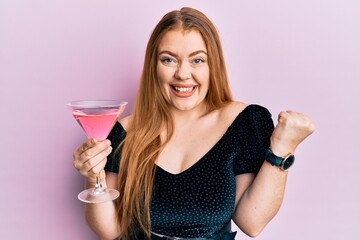 Young beautiful redhead woman drinking tropical cocktail with alcohol screaming proud, celebrating victory and success very excited with raised arm