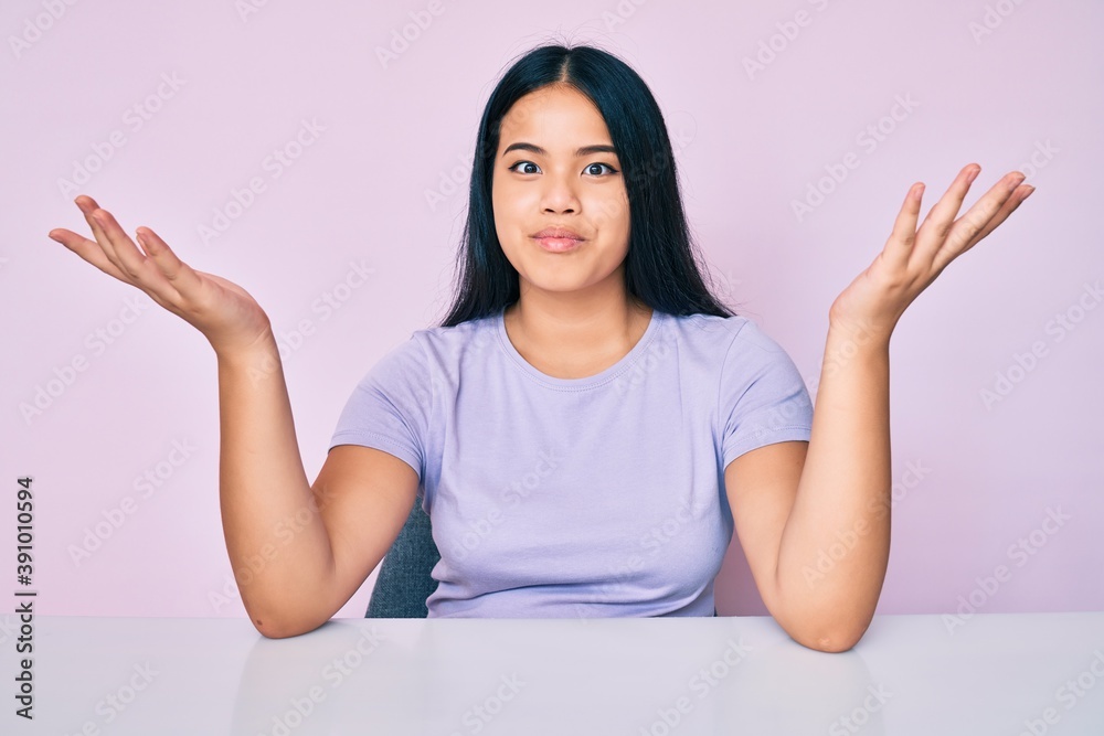 Young beautiful asian girl wearing casual clothes sitting on the table clueless and confused expression with arms and hands raised. doubt concept.